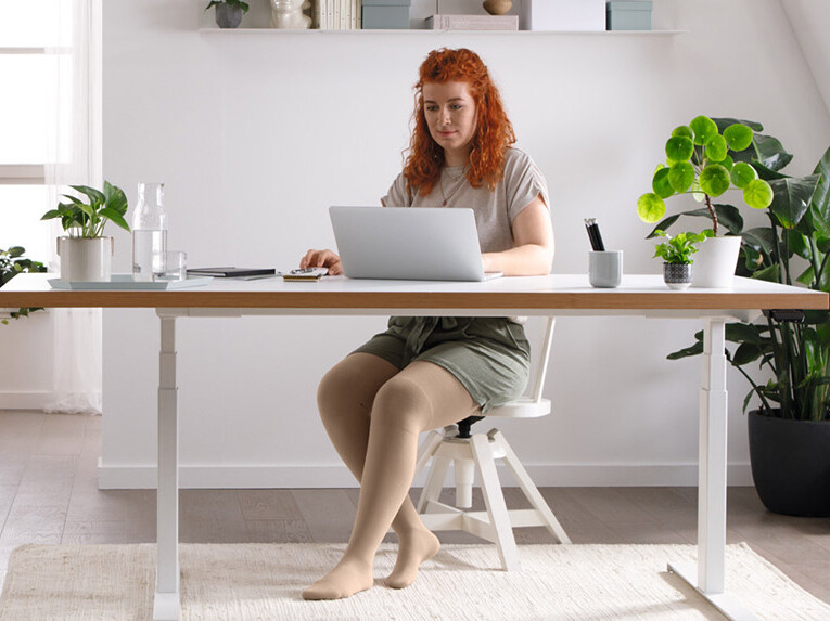 Wissenswertes über Lipödem Eine Frau sitzt an einem Schreibtisch an ihrem Laptop, sie trägt Juzo Expert Flachstrickkompressionsstrümpfe