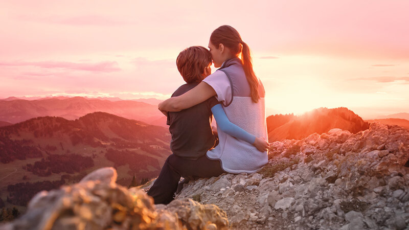 A mother is sitting on a mountain peak with her child. The child is wearing a compression sleeve. A mother is sitting on a mountain peak with her child. The child is wearing a compression sleeve.