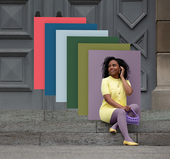 Uma mulher está sentada nos degraus de pedra em frente a um portão. Ela usa um conjunto de duas peças amarelo claro com uma compressa roxa. Atrás dela, podem-se ver campos de cores nas cores da moda