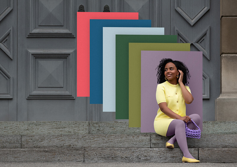 Juzo Trend Colours Batik En kvinde sidder på stentrapper foran en port. Hun bærer et lysegult todelt sæt til en lilla kompressionsbandage. Bag hende ses farvefelter i trendfarverne.