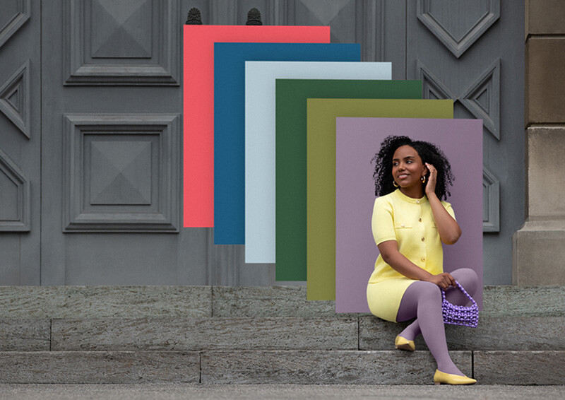 Trend Colours Batik de Juzo Una mujer está sentada en unos escalones de piedra frente a una puerta. Lleva un conjunto de dos piezas de color amarillo claro y una compresión de color lila. Detrás de ella se ven campos de color en los colores de moda.