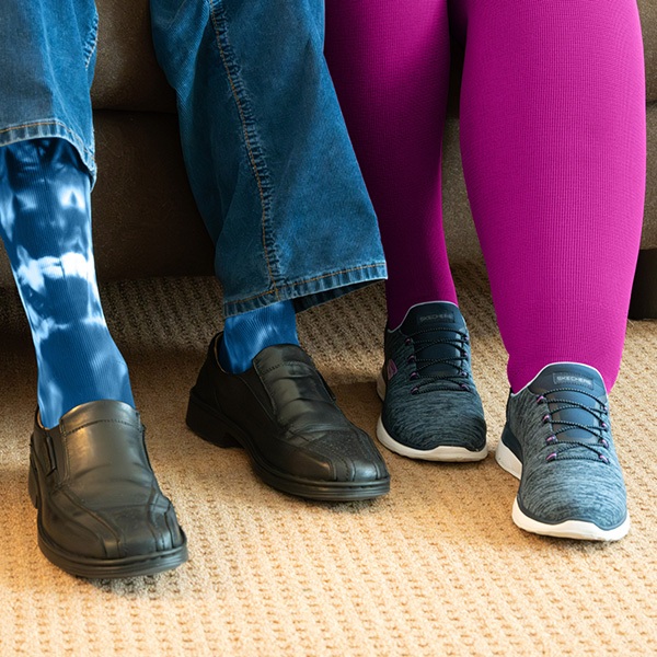 Man sieht die Beine von einem Mann mit einer Kompressionsversorgung, die mit Batik gefärbt wurde. Daneben sieht man die Beine einer Dame mit einer pinken Kompressionsversorgung.