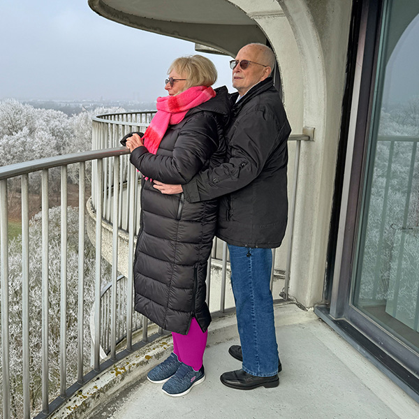 Ein Mann umarmt eine Frau mit Lymphödem auf einem Balkon, ihr Blick schweift in die Ferne