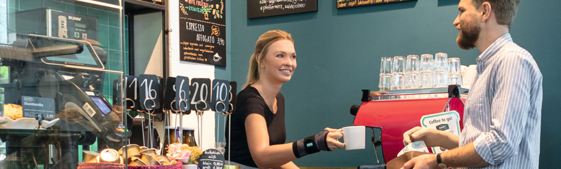 Lifestyle-afbeelding Café - JuzoFlex Manu Xtra Verkoopster in bakkerij draagt JuzoFlex Manu Xtra polsbandage en reikt klanten over de toonbank een kopje aan