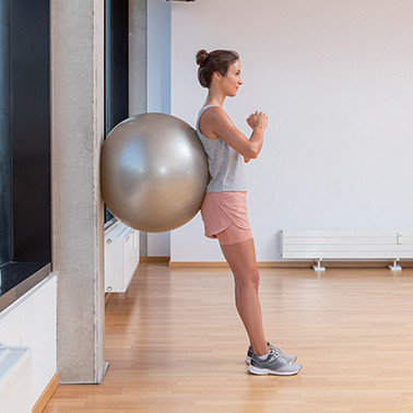Uebung Wall Sit Schritt 1 Frau lehnt sich aufrecht mit Gymnastikball an eine Wand