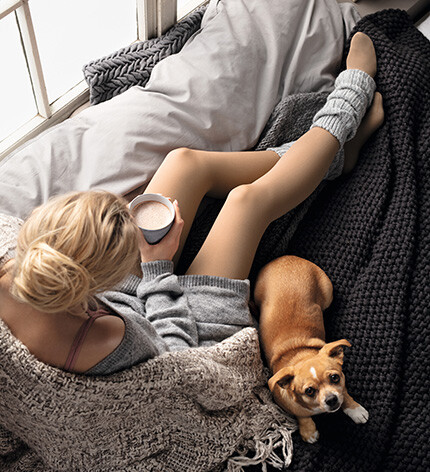 Eine Frau sitzt in eine Decke gekuschelt auf der Couch und trinkt einen Kakao